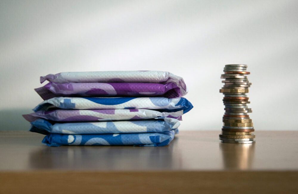 A stack of sanitary towels next to a taller stack of coins. Illustrating period poverty and unaffordability of sanitary materials for many women worldwide.
