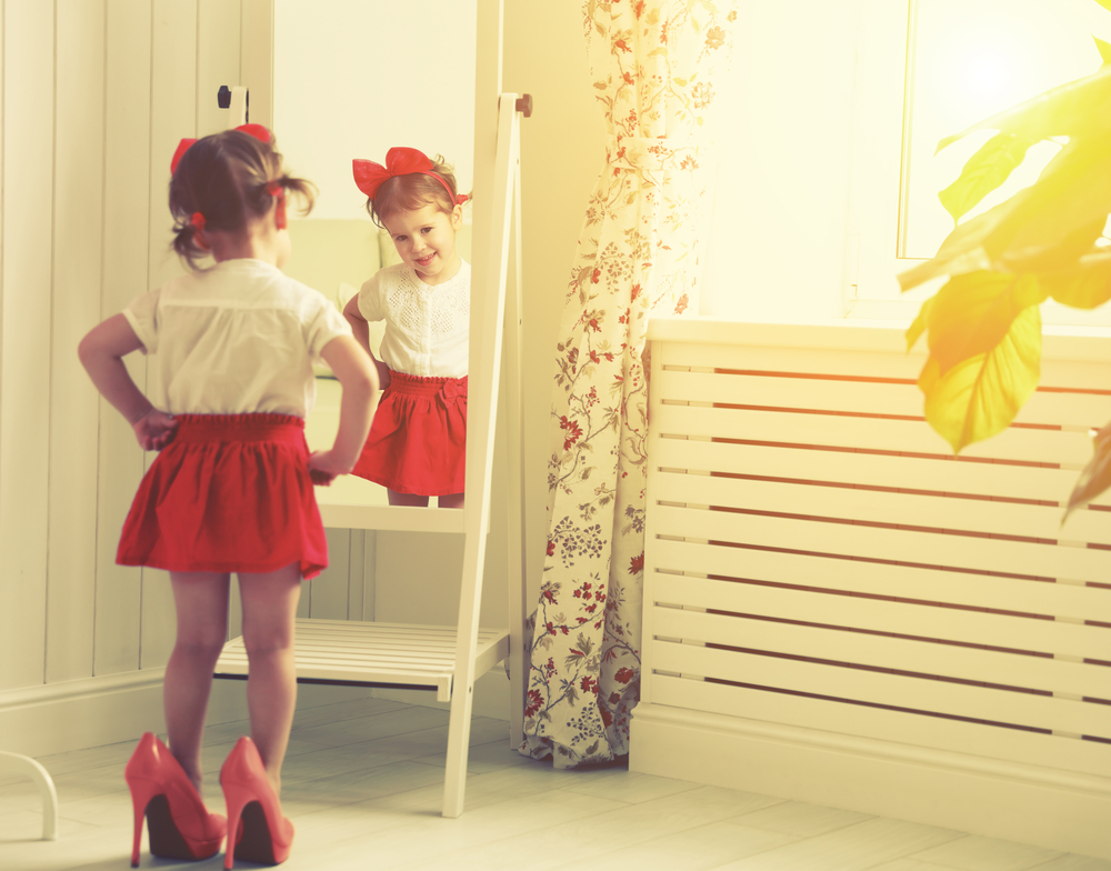 little girl child fashionista looking in the mirror at home in a red skirt, shoes of mother