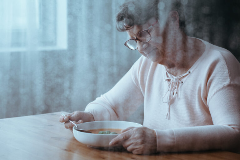 An elderly woman sits at a table over a bowl of soup, looking sullen and depressed